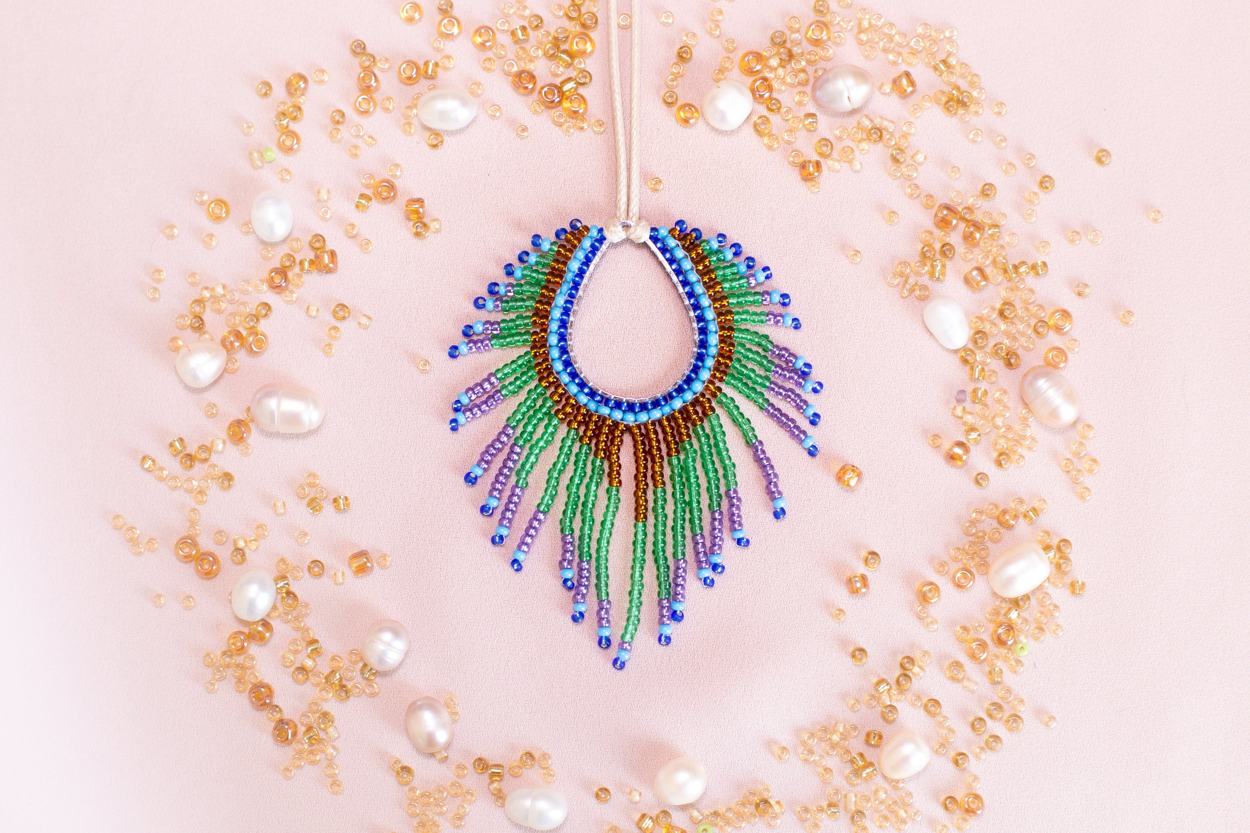 a colorful beaded necklace with a large, eye-catching pendant surrounded by a variety of small, round, and multicolored beads, pearls, and other decorative elements arranged in a circular pattern on a pink background.